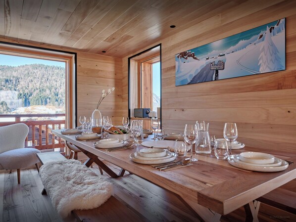 A wooden dining table set with plates, glasses, and cutlery, overlooking a snowy mountain landscape.