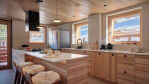 A modern kitchen with wooden cabinets, a central island, bar stools, and a large window with a view of snow-capped mountains.