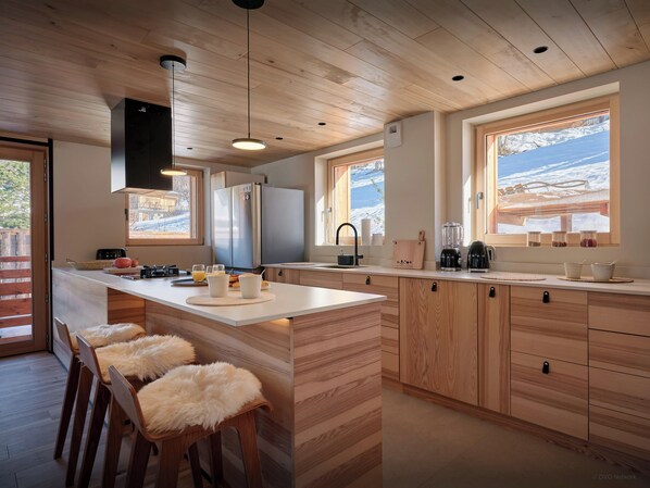 Une cuisine moderne avec des meubles en bois, un îlot central, des tabourets de bar et une grande fenêtre offrant une vue sur des montagnes enneigées.