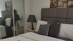 Ein ordentlich bezogenes Bett mit weißer Daunendecke und einem gefalteten grauen Handtuch. Ein Nachttisch mit Lampe und ein Gemälde einer Stadtansicht an der Wand.