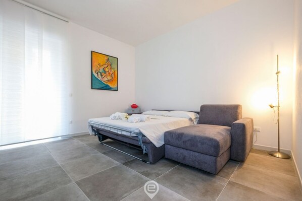 A modern bedroom with a bed, a grey sofa, a floor lamp, and a painting on the wall.