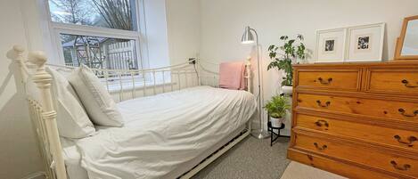 A bedroom with a single bed, a dresser, a floor lamp, a potted plant, and framed pictures on the wall.