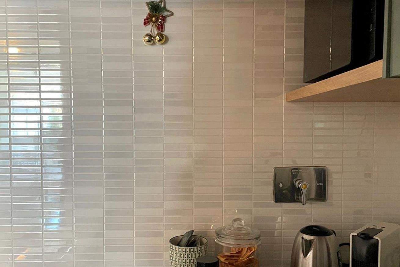 A kitchen with white tiled walls, a stainless steel kettle, a coffee maker, and a small cabinet.