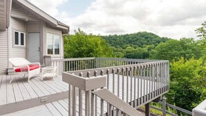 Une terrasse en bois avec une balustrade blanche, une chaise blanche avec un coussin rouge et une fenêtre donnant sur une colline boisée.