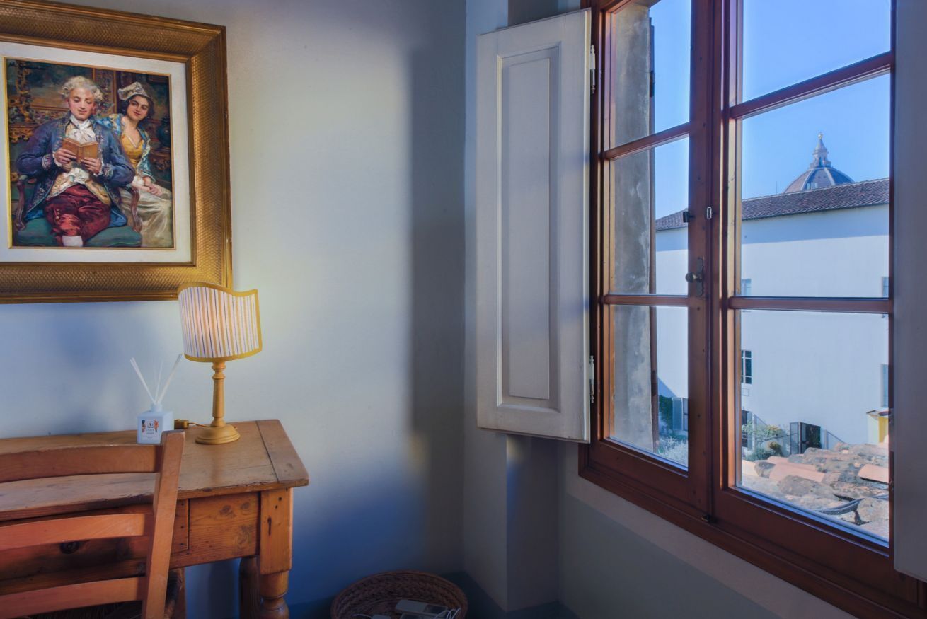 A room with a wooden table, a lamp, a framed painting, and a window with an open shutter.