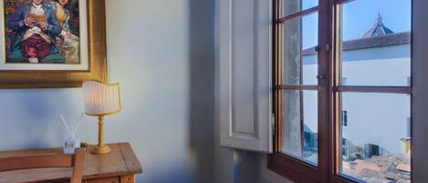 A room with a wooden table, a lamp, a framed painting, and a window with an open shutter.