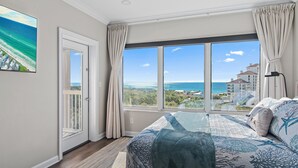 A bedroom with a large window offering a view of the ocean and buildings.