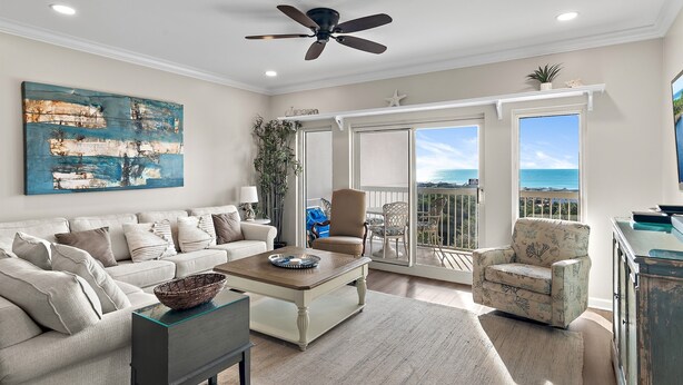 A living room with a sofa, coffee table, and armchair, a ceiling fan, and a view of the ocean through large sliding doors.