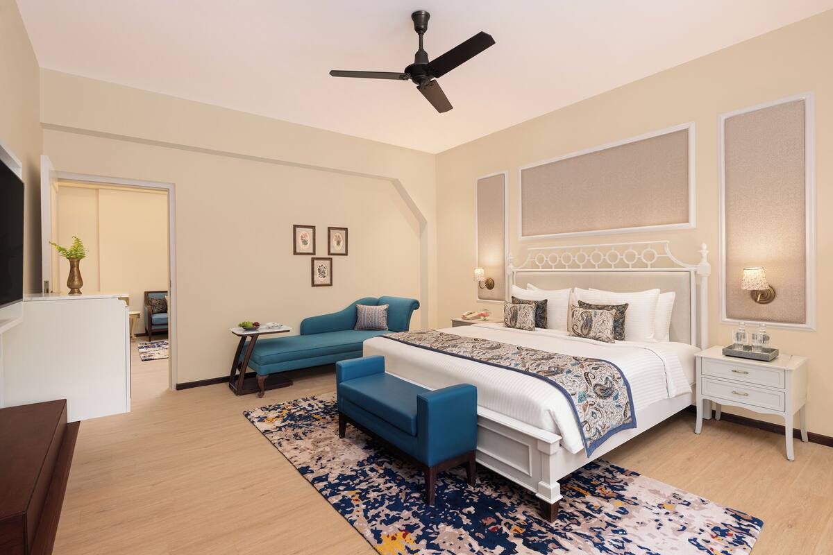 A bedroom with a bed, a blue sofa, a white bedside table, a ceiling fan, and a large window.