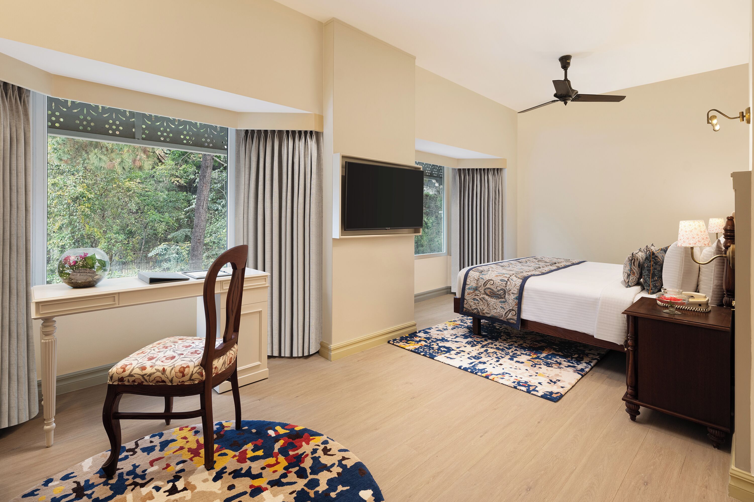 A bedroom with a bed, a desk, a chair, a TV, and a view of trees through the window.
