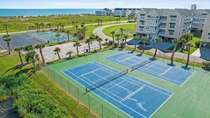 Complejo de canchas de tenis con varias canchas, rodeado de un estacionamiento y palmeras, junto a un edificio con balcones.