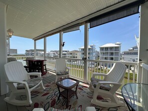 A covered outdoor area with white chairs, a glass table, and a view of beachfront buildings.