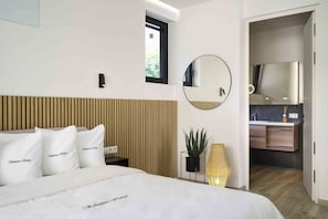 A modern bedroom with a wooden headboard, a round mirror, a bedside table with a lamp, and a view into the bathroom. - SalvatoreHomes - Pure Luxury in the Centre of Bardolino with Pool & Private Jacuzzi - Casa Olivia - a 5 Star Luxury Collection at Lake Garda (Bardolino)