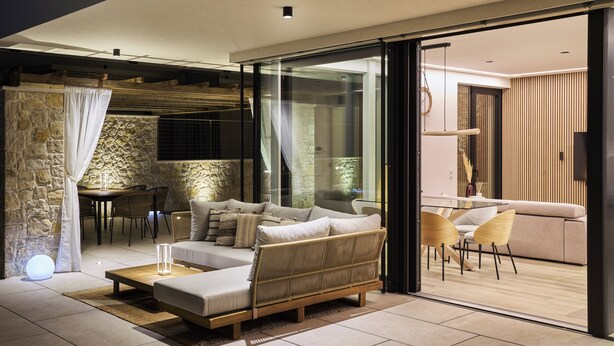 A modern living room with a sofa, coffee table, and chairs. There is a glass door leading to a dining area with a table and chairs.