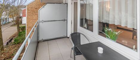 Un balcon avec une table et une chaise, une porte blanche et une vue sur un jardin et des bâtiments.