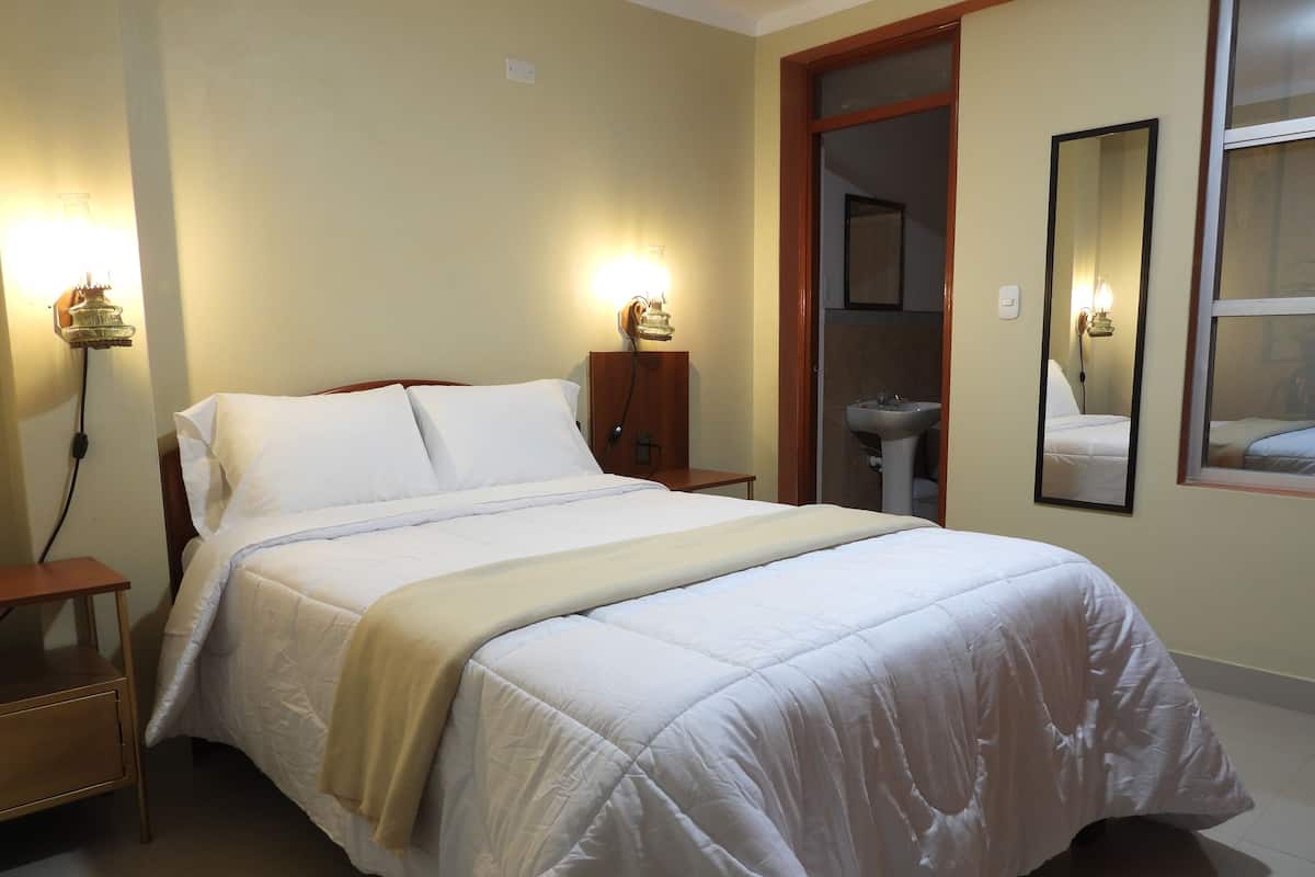 Un lit bien fait, avec du linge de lit blanc, une table de chevet, une commode, une lampe fixée au mur et un miroir.