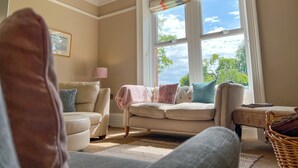 A cozy living room with a beige sofa, a small armchair, and a wicker chair. A large window with a view of trees and a clear sky is visible. - High House, Southwaite -  a cottage that sleeps 10 guests  in 5 bedrooms (Southwaite)