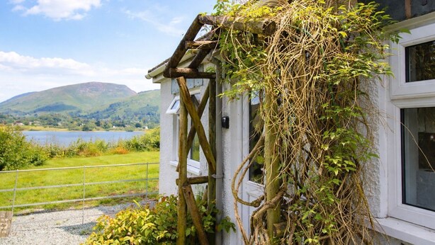 A rustic building with a window, a wooden structure attached to the wall, and a view of a lake and hills in the background.