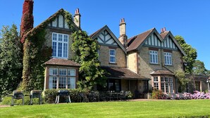 Een groot, historisch huis met meerdere schoorstenen, een verzorgd gazon en een patio met zitplaatsen buiten.