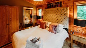 Ein Schlafzimmer einer Holzhütte mit einem großen Bett, einem Spiegel, einem Schild mit der Aufschrift 'NEED IS LOVE' und einem Fenster mit Blick auf Bäume.