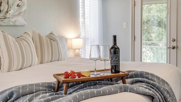 Una cama impecablemente arreglada con una bandeja de fresas, una botella de vino y copas, todo sobre un pared azul claro con una ventana y cortinas.
