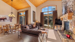 A spacious living room with a stone fireplace, leather sofas, a dining area with wooden chairs, and large windows offering a view of the outdoors.