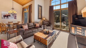 A cozy living room with a leather sofa, a coffee table, a TV, and a large window offering a view of the outdoors.