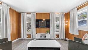 Una sala de estar moderna con televisor de pantalla plana, paneles de madera, una consola blanca y un gran ventanal con cortinas.