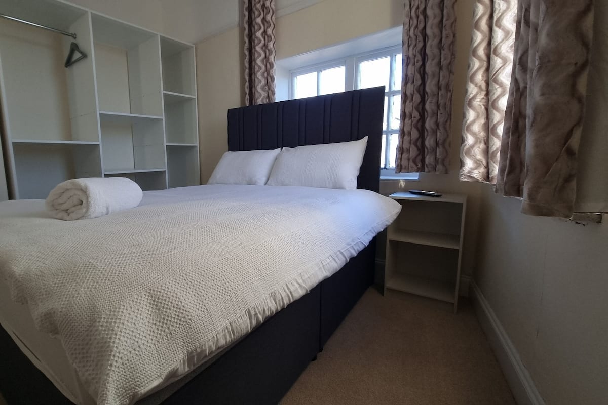 A neatly made bed with a white textured blanket and a rolled towel, a dark headboard, and a bedside table with a remote control.