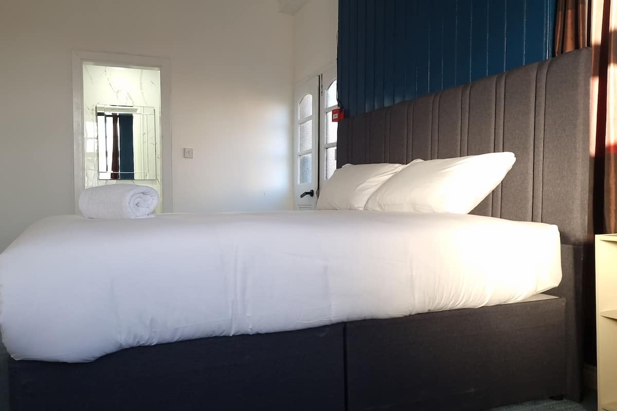A neatly made bed with white linens in a room with a dark headboard and a window with curtains.