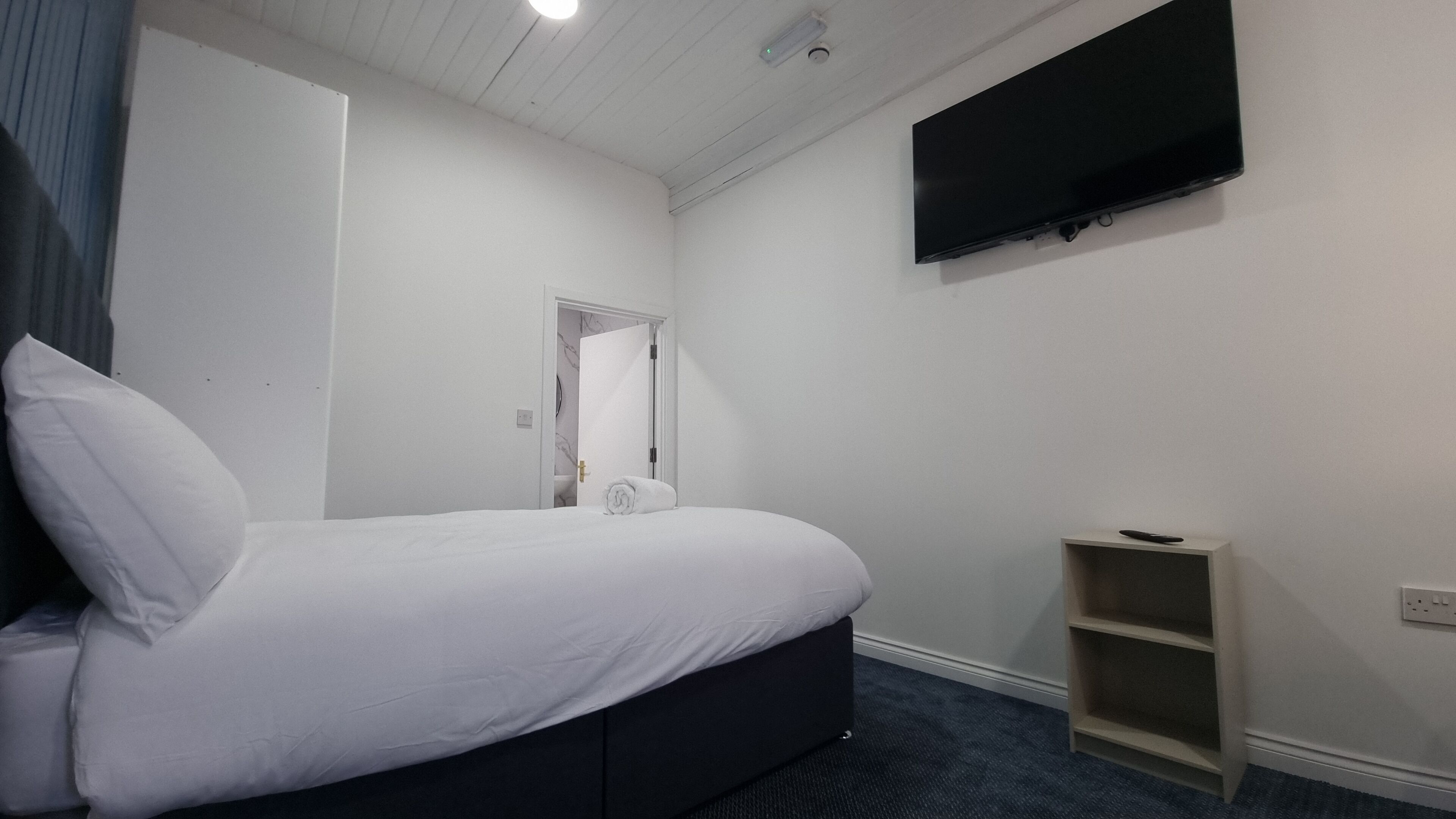 Un dormitorio con cama, televisor montado en la pared y un pequeño estante de madera.