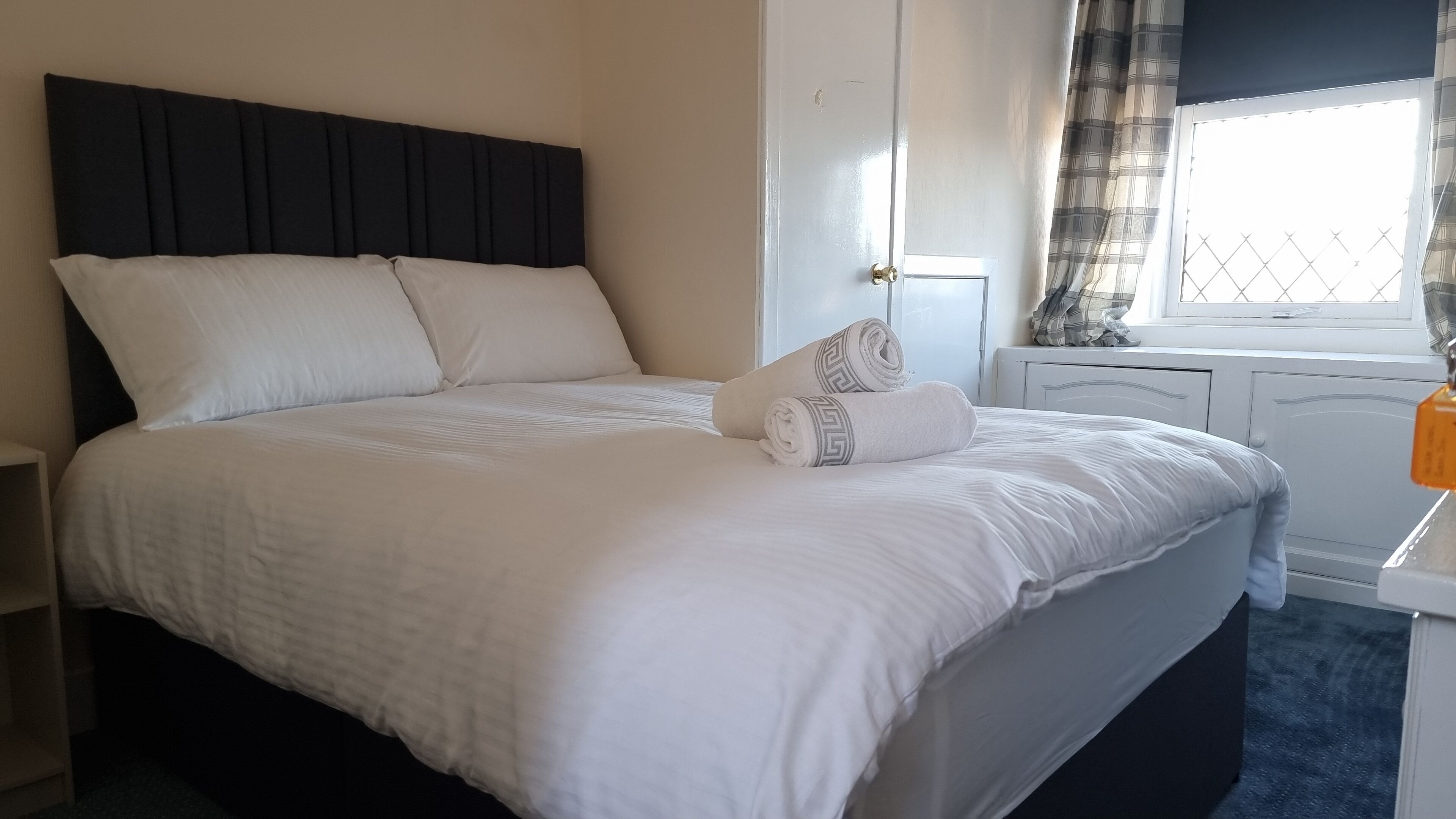 A neatly made bed with white linens and a dark headboard, a window with curtains, and a small bedside table with a lamp.