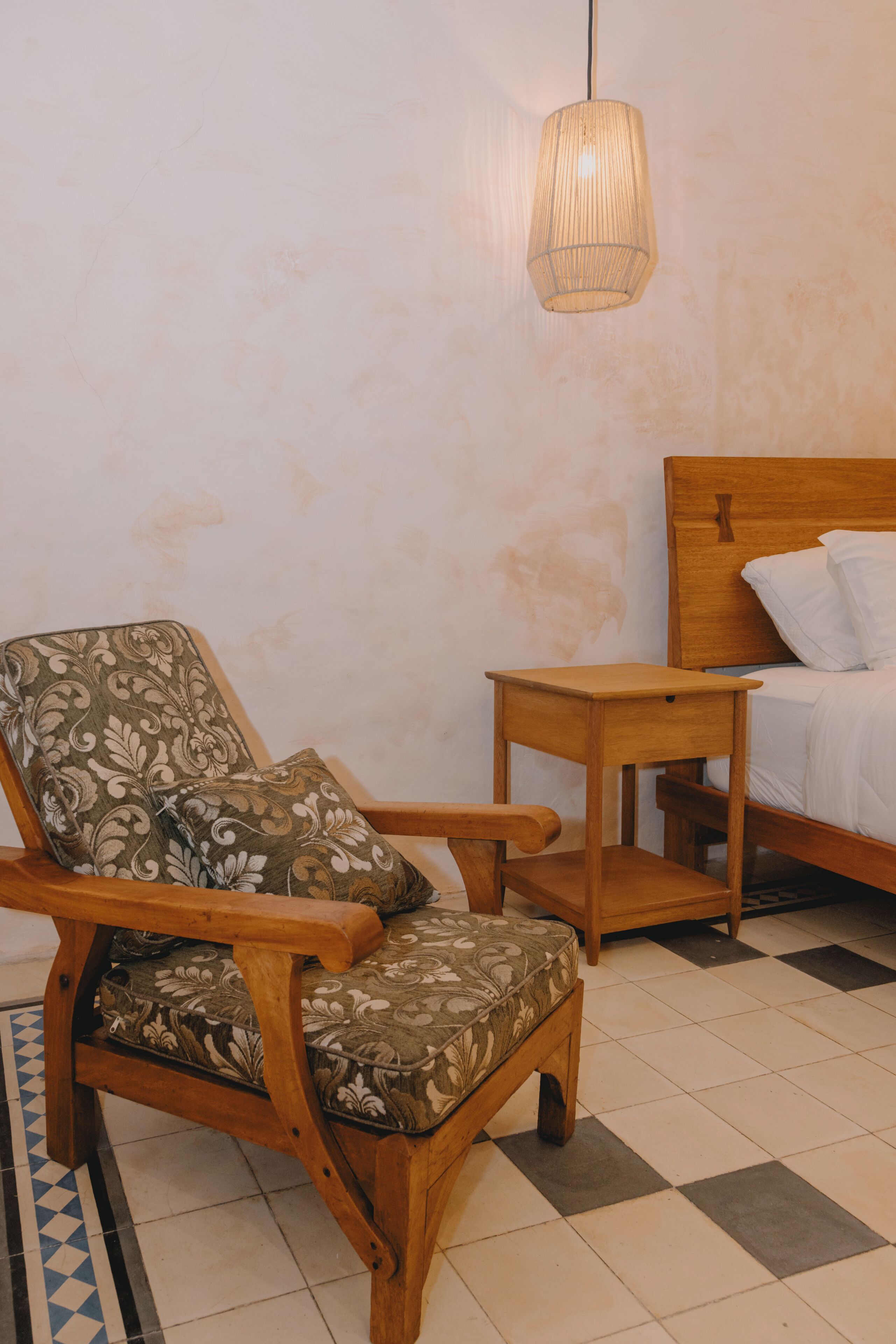 Una silla de madera con tapicería floral, una mesita de noche de madera y una cama con ropa de cama blanca.