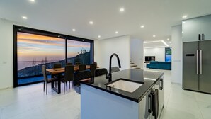 Een moderne keuken met een groot eiland, een eethoek met uitzicht en een woonruimte met televisie en bank.