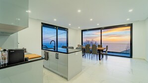 Een moderne keuken met een inbouwfornuis, een eethoek met een tafel en stoelen, en grote ramen met uitzicht op de zee bij zonsondergang.