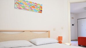Un dormitorio con cabecera de madera, ropa de cama blanca, un cuadro de peces de colores, una silla roja y una puerta que da a otra habitación.