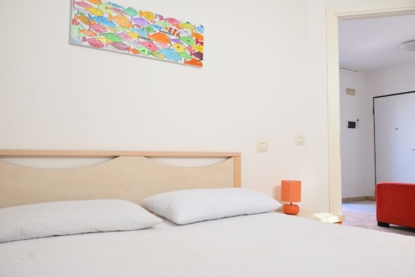Une chambre avec une tête de lit en bois, du linge de lit blanc, une décoration murale représentant des poissons aux couleurs vives, une chaise rouge et une porte donnant sur une autre pièce.