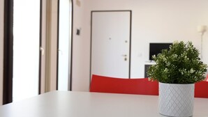 Una habitación con una mesa blanca, una silla roja, una maceta blanca con textura y una puerta cerrada al fondo.