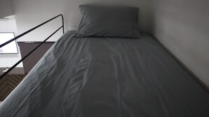 Un lit simple avec un matelas et un oreiller gris, dans une pièce aux murs blancs et au sol marron.