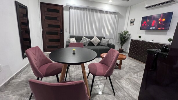 Un salon moderne avec une table à manger ronde, deux chaises roses, un canapé gris agrémenté de coussins à motifs, un téléviseur fixé au mur et une plante en pot.