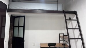 Un lit superposé avec une échelle, un bureau et une chaise, dans une pièce aux murs blancs et dotée d’une porte à panneaux de verre.
