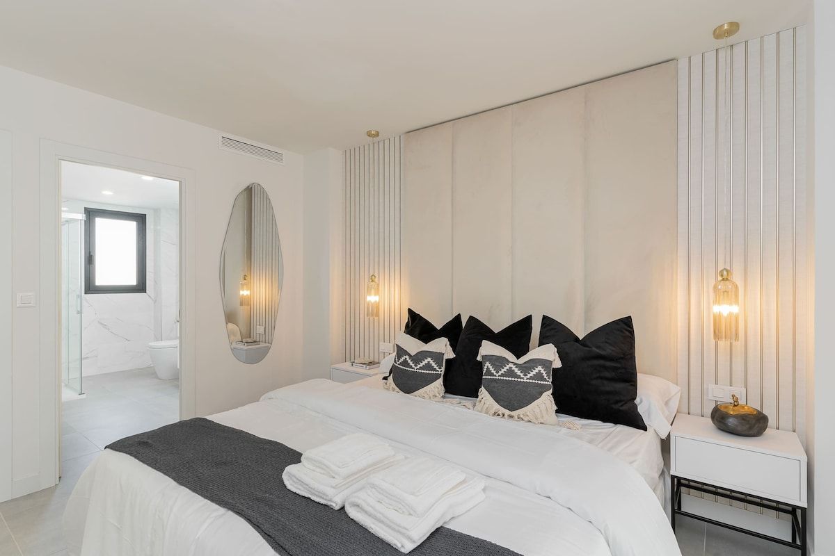 A modern bedroom with a large bed, bedside lamps, a mirror, and a view of the bathroom.