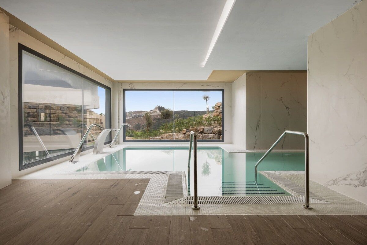 A modern indoor swimming pool with clear glass walls, a view of rocky terrain, and a wooden floor.