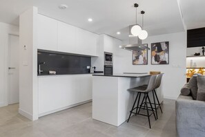 A modern kitchen with white cabinetry, a central island with bar stools, and a dining area with a sofa. - La Brisa | Pool - terrace - gym (Estepona)