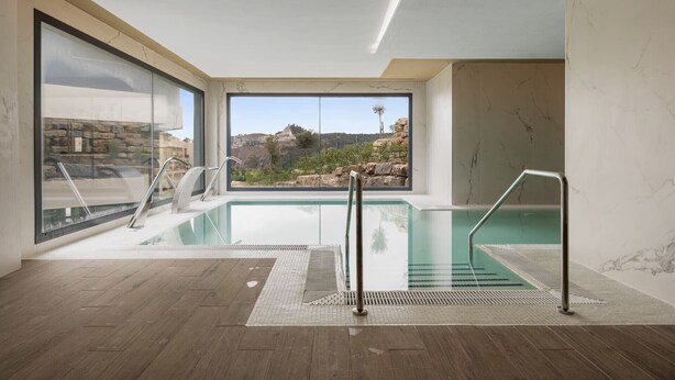 A modern indoor swimming pool with clear glass walls, a wooden floor, and a view of rocky terrain outside. - La Brisa | Pool - terrace - gym (Estepona)