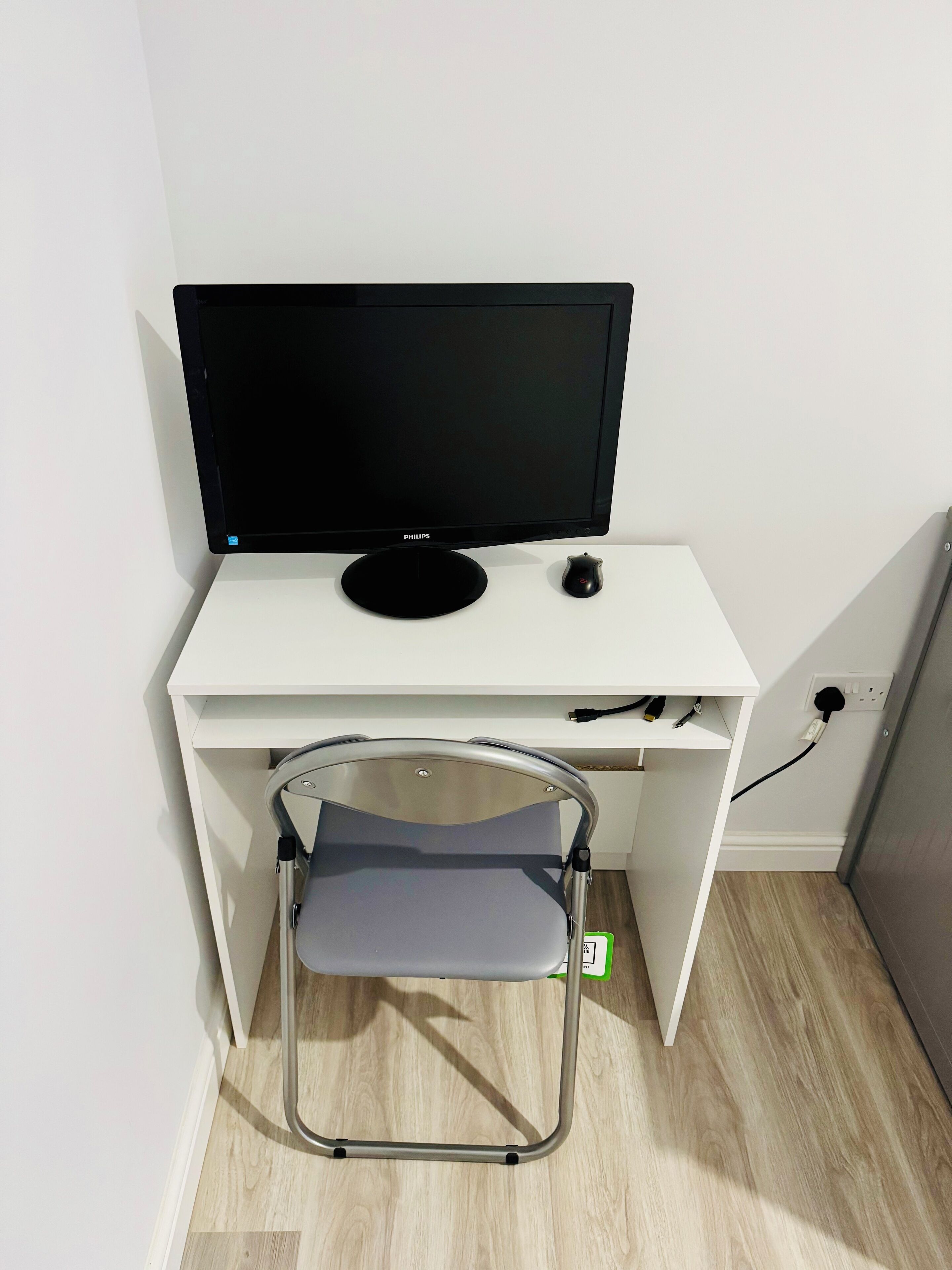 Een wit bureau met een zwart beeldscherm, een klein zwart voorwerp en een opklapbare stoel.