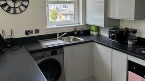 A modern kitchen with white cabinets, a black countertop, a built-in washing machine, a window with a view, and a clock on the wall. - Glass Street Apartment (Glenrothes)