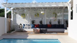A poolside area with a white pergola, hanging plants, a blue sofa, and a wicker basket.