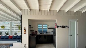 A modern kitchen with a built-in oven, a sink, and a window with blinds. There is a sign on the wall that reads "POOL TOWELS".