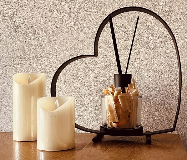 Tres velas sobre una mesa de madera con un candelabro en forma de corazón que contiene un difusor con flores secas.